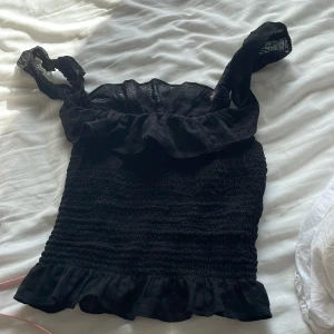 Svart volangtopp från ASOS - Supersöt svart topp från ASOS med smockad kropp och volangdetaljer både upptill och nertill. Axelbandsmodell med breda axelband och lätt transparent tyg upptill. Perfekt för dig som gillar en feminin och trendig look.