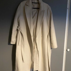Beige lång kappa med bälte - Snygg beige kappa i lång modell med bälte i midjan och klassisk krage. Jackan har knappar framtill och raka ärmar. Perfekt för dig som vill ha en stilren och tidlös look under höst och vinter.
