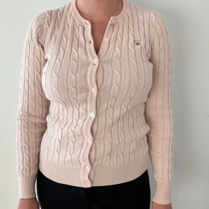 Babyrosa kabelstickad kofta från GANT - Säljer en klassisk babyrosa kabelstickad kofta från GANT med rund halsringning och knappar framtill. Koftan har lång ärm och broderad logga på bröstet. Perfekt för lager-på-lager och stilren vardagslook. Färg: babyrosa