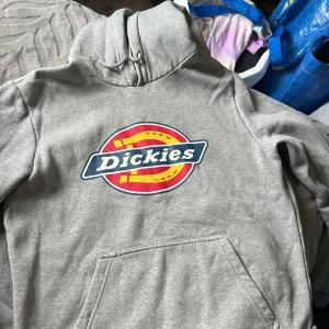 Grå Dickies hoodie med logga - Säljer en grå hoodie från Dickies i storlek XS. Tröjan har stor färgglad Dickies-logga på bröstet, huva med snörning och en klassisk magficka. Perfekt för dig som gillar streetwear och vill ha en enkel men snygg look.