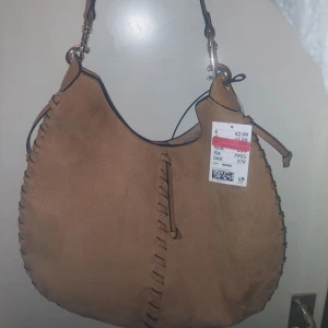 Hel ny H&M -  Brun axelväska i fejk mocka - Säljer en snygg beige axelväska med mjuk mocka-känsla och coola flätade detaljer längs kanterna. Väskan har en bred axelrem med silverfärgade metalldetaljer och en avslappnad, rundad form. Perfekt för dig som gillar boho-vibes och vill ha plats för allt du behöver. Den har ingen dragkedja utan bara knapp för att stänga väskan.
