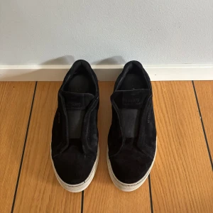 Svarta sneakers i mocka från Axel Arigato - Stilrena svarta sneakers från Axel Arigato i mjuk mocka med vit platt sula. Skorna har minimalistisk design utan snörning och diskret logga på sidan. Perfekta för dig som gillar en clean och modern look. Du får med lådan och allt vad den inehåller. Storlek 42 dock små i storleken. Priset är ej fast och kan ändras vid snabb affär. 