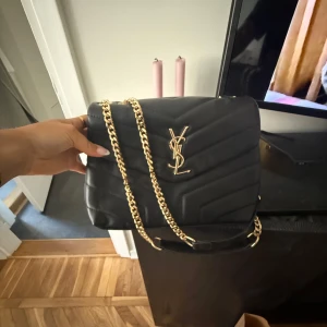 Svart quiltad YSL axelväska i skinn - Säljer en svart quiltad axelväska från YSL med ikoniskt guldigt YSL-emblem och kedjerem. Väskan är tillverkad i mjukt skinn och har en stilren, lyxig look med snyggt mönstrad yta. Perfekt för dig som vill ha en statementväska med exklusiv känsla. (Ej äkta) som ny