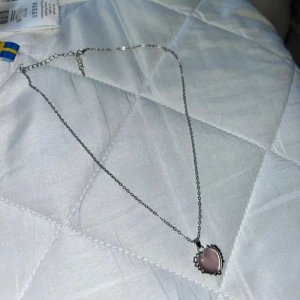Halsband med rosa hjärthänge - Snyggt halsband med tunn silverfärgad kedja och ett hjärtformat hänge i ljusrosa. Hänget har en dekorativ kant som ger en cool och unik look. Perfekt för dig som gillar söta detaljer och vill ha något som sticker ut.