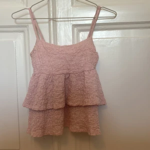 Rosa volangtopp från Gina Tricot XS - Supersöt ljusrosa linnetopp från Gina Tricot i storlek XS. Toppen har smala axelband, volanger i två lager och ett krinklat tyg som ger en cool struktur. Perfekt för varma dagar när du vill ha något luftigt och trendigt.den har justerbara axelband och jag säljer för 90kr