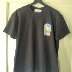 Svart Casablanca t-shirt med tryck - Svart t-shirt från Casablanca Tennis Club med färgstarkt grafiskt tryck fram och bak i blått, orange, gult och rosa. Rund hals, korta ärmar och normal passform. Materialet är mjuk bomull och trycket har en cool, modern vibe. Den ända defekten ser ni på sista bilden, men prislappen sitter fortfarande kvar!