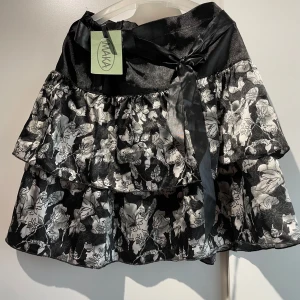 Blommig volangkjol med rosett - Supersöt kort volangkjol från Maka i svart och silver med blommigt mönster. Har aldrig används💞