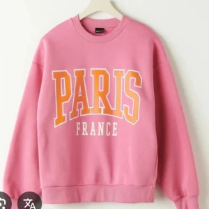 Rosa sweatshirt med Paris-tryck - Säljer en rosa sweatshirt med stort tryck 'PARIS FRANCE' i orange och vitt på bröstet. Tröjan har rund hals, långa ärmar och en loose passform. Perfekt för dig som gillar streetstyle och vill sticka ut med färg.