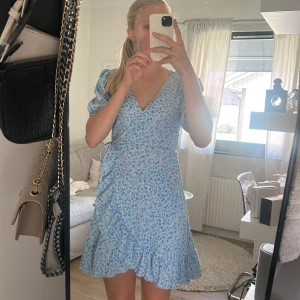 Blommig klänning i ljusblått - Superfin kort omlottklänning med ljusblått blommigt mönster och volangkant längst ner. Klänningen har puffiga korta ärmar och knyt i midjan för en snygg passform. Perfekt för sommaren! Köpt för 400 kr.