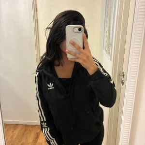 Svart Adidas vindjacka med huva - Svart vindjacka från Adidas med klassiska vita ränder längs ärmarna och broderad logga på bröstet och ärmen. Jackan har huva, dragkedja framtill och två fickor. Aldrig använd. 