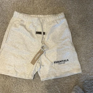 Grå Essentials Fear of God shorts - Snygga ljusgrå shorts från Essentials Fear of God med svart logotyptryck på benet och patch framtill. De har elastisk midja med snörning och är gjorda i mjuk bomull, perfekt för chill dagar. Klassisk loose passform och stilren look.