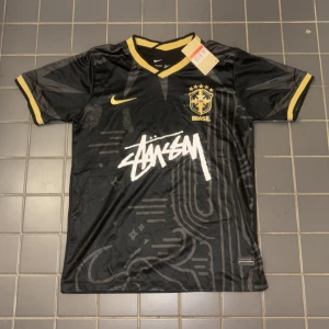Nike x Stüssy Brasil svart fotbollströja - Limited edition svart Brasilien fotbollströja från Nike och Stüssy. Tröjan har gula detaljer på krage och ärmslut, CBF-logga på bröstet och Stüssy-tryck framtill. Tillverkad i Dri-FIT-material som andas, perfekt för fotboll eller streetwear.