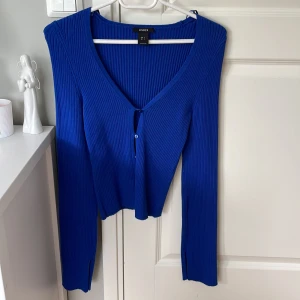 Blå ribbad kofta från Lindex - Snygg blå kofta från Lindex med ribbad struktur och v-ringning. Modellen har långa ärmar, knappar framtill och en figursydd passform som ger en trendig look. Perfekt att styla med jeans eller kjol för en cool vardagsoutfit.