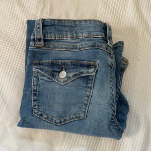 Blå bootcut jeans från Gina Tricot - Snygga blå jeans från Gina Tricot i bootcut-modell med slitna detaljer på knäna och klassiska fickor med knapp bak. Jeansen har normal passform och är tillverkade i jeansmaterial med ljus tvätt och kontrastsömmar. Perfekt för dig som gillar en avslappnad och trendig look.