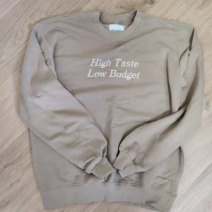 Beige sweatshirt från Yaliny - Säljer en beige sweatshirt från Yaliny med trycket 'High Taste Low Budget' i vitt på bröstet. Tröjan har rund hals, långa ärmar och ribbade muddar vid ärmslut och nederkant. Perfekt för chill dagar och streetstyle.