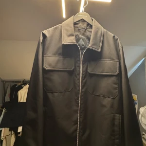 Svart overshirt från Moose Knuckles (med kvitto) - Helt ny snygg svart overshirt från Moose Knuckles med raka linjer, stora bröstfickor och dragkedja framtill. Jackan har diskret logga på ärmen och innerfickor med dragkedja. Perfekt för dig som gillar stilren och clean look. Köpt från Boozt.com och kvittto finns. 