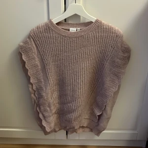 Stickad volangtröja från Åhléns  - Supersöt stickad tröja i ljuslila nyans. Tröjan är ärmlös med breda volangdetaljer längs sidorna och har en grovstickad struktur🩷