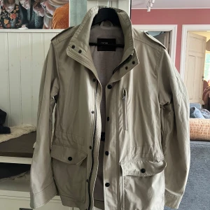 Beige vindjacka field jacket från Oscar - Snygg beige vindjacka från Oscar Jacobsson med hög krage, flera fickor med knappar och dragkedja framtill. Jackan har en clean look och är perfekt för dig som gillar stilrena plagg. Passar till många outfits och har lång ärm.