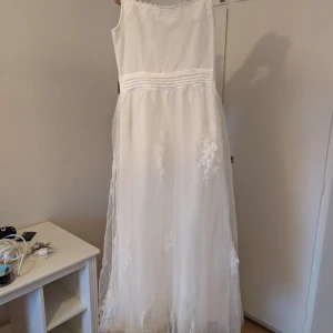 Vit långklänning med spetsdetaljer - Säljer en elegant vit långklänning med tunna axelband och vacker spetskant upptill. Klänningen har flera lager tunn tyll och fina broderade blommor på kjolen. Midjan är markerad med diskreta veck och baktill finns en draperad detalj. Perfekt för speciella tillfällen!