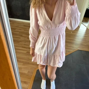Zara Ljusrosa kort klänning med volang - Supersöt ljusrosa klänning med v-ringning och smock i midjan och vid ärmslut. Klänningen har långa ärmar och volangkant nedtill som ger en romantisk vibe. Perfekt för dig som gillar en feminin och trendig stil. Storlek M men passar mig S