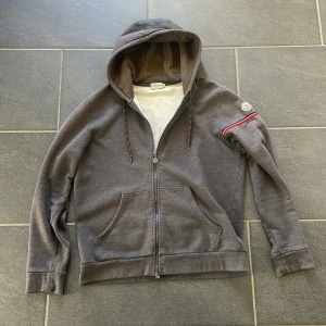 Grå Moncler hoodie med dragkedja - Snygg grå hoodie från Moncler med huva och hel dragkedja framtill. Tröjan har Moncler-logga på ärmen. Står att den är Xl men passar mer som M