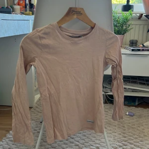 Ljusrosa långärmad tröja 157 Denim Makers - Ljusrosa långärmad tröja från 157 Denim Makers med rund hals och diskret logotyp-patch nedtill. Tröjan är i mjuk bomull och har en enkel, clean look som funkar till jeans eller kjol. Perfekt för dig som gillar basic och comfy vibes.