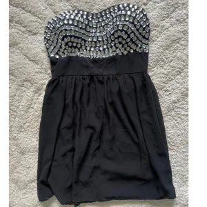 Svart axelbandslös klänning strass - Snygg svart axelbandslös kortklänning från Oneness med glittrig strass upptill. Klänningen är gjord i 100% polyester och har en lätt utställd kjol. Perfekt för dig som vill sticka ut med lite extra bling.Axellös bandlös tubklänning Klänning från oneness svart med ”diamanter”. Använd 1 gång, mycket bra skick. Storlek L. Går strax under rumpan på mig som är 168