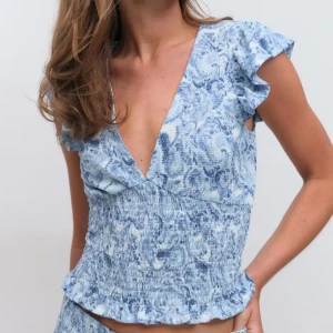 Blå blommig blus med volang - Supersöt blommig blus i ljusblå toner med djup v-ringning och volangärmar. Smockad midja och volangkant nedtill ger en feminin och trendig look. Perfekt för varma dagar och somriga vibes.
