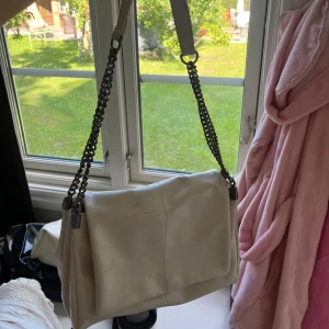 Beige axelväska med kedjedetaljer - Snygg beige axelväska i skinnimitation med bred axelrem och coola kedjor i metall. Väskan har en stilren, fyrkantig form och stora synliga sömmar som ger en modern vibe. Perfekt storlek för att få plats med allt du behöver till vardags.