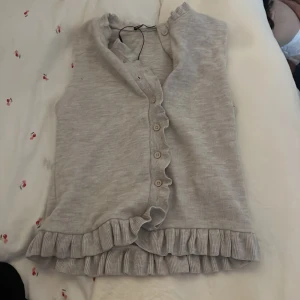Grå volangväst från Zara - Supersöt grå väst från Zara med volangdetaljer längs knäppningen och nederkanten. Västen är ärmlös, har knappar framtill och är gjord i ett mjukt, ribbat material som känns riktigt comfy och trendigt. Liten i storlek 💖💖