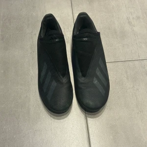 Svarta fotbollsskor från Adidas - Säljer ett par svarta fotbollsskor från Adidas med en stilren design och diskreta ränder. Skorna har en strömlinjeformad form utan snörning och är perfekta för spel på gräsplaner. De har en robust sula med dobbar för optimalt grepp. Lite slitna i sulorna.