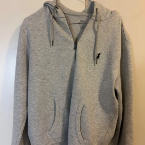 Grå hoodie från Ralph Lauren - Säljer en stilren grå hoodie från Ralph Lauren med dragkedja och klassisk logga på bröstet. Tröjan har en bekväm passform och är perfekt för kyligare dagar. Den har en praktisk känguruficka och justerbar huva. Perfekt för en avslappnad look. 9/10 skick. 