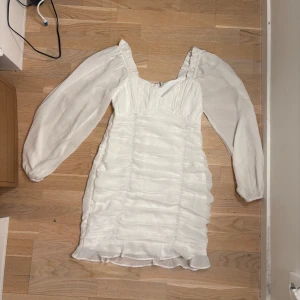 Vit klänning från NLY Trend - Säljer en elegant vit klänning från NLY Trend med långa puffärmar och dragkedja i ryggen. Klänningen har en vacker struktur med volanger och är perfekt för speciella tillfällen. 