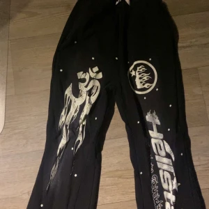 Hellstar Sweatpants - Coola svarta byxor med unika tryck i vitt, inklusive text och symboler. Byxorna har en avslappnad passform och elastisk midja för extra komfort. Perfekta för en avslappnad stil med en twist.