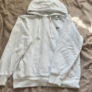 Lacoste hoodie - Säljer en stilren vit hoodie från Lacoste Sport med den klassiska krokodilloggan på bröstet. Hoodien har en justerbar huva med snören och en bekväm passform. Perfekt för en avslappnad look.