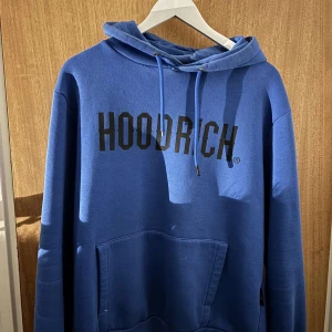 Blå hoodie från Hoodrich - Säljer en snygg blå hoodie från Hoodrich med svart text på bröstet. Den har en klassisk känguruficka och justerbar huva med snören. 