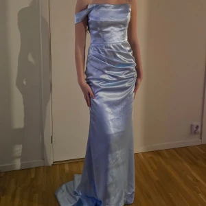 Ljusblå axelbandslös långklänning - Elegant ljusblå långklänning i satin med offshoulder-design. Klänningen har en smickrande passform och ett vackert fall som ger en lyxig känsla. Perfekt för speciella tillfällen.