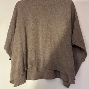 Beige stickad poncho softgoat  - Säljer en mysig beige stickad poncho med en lös passform. Perfekt för kyliga dagar och ger en avslappnad stil. Den har en enkel design med ribbade kanter.
