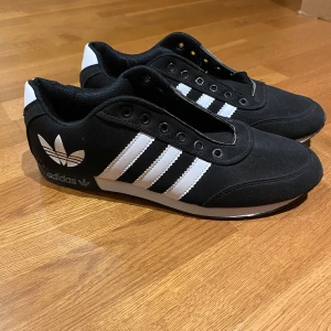 Svarta och vita sneakers från Adidas - Snygga svarta sneakers med vita detaljer från Adidas. Skorna har den klassiska tre-randiga designen på sidan och en mönstrad sula för bra grepp. Perfekta för en stilren look.
