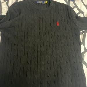 Svart kabelstickad tröja från Polo Ralph Lauren - Säljer en stilren svart kabelstickad tröja från Polo Ralph Lauren. Tröjan har en klassisk design med rund hals och det ikoniska röda broderade logotypen på bröstet. Perfekt för en elegant och avslappnad look.
