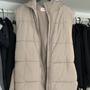 Beige dunväst med luva - Snygg beige dunväst med huva och dragkedja framtill. Västen har ett bälte i midjan för en justerbar passform. Perfekt för kyligare dagar.