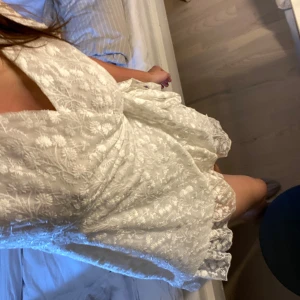 Vit  klänning - Säljer en söt vit klänning med blommigt mönster och korta ärmar. Klänningen har en omlott-design och volangdetaljer vid ärmslut och nederkant. Perfekt för sommarens alla tillfällen! Använd 1 gång vid ett studentfirande, kan användas till student eller som en sommarklänning. Klänningen är som ny. Klänningen är i storlek L men skulle säga att den även passar de som är M eftersom den går att dra åt hur mycket man vill. 