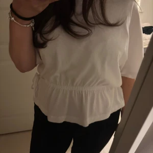 Vit kortärmad topp från H&M - Säljer en stilren vit topp från H&M med korta ärmar och en elastisk midja med knytband. Perfekt för en avslappnad look.