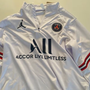 PSG fotbolls tröja  - Säljer en vit PSG fotbollströja med lång ärm och Jordan-logga. Tröjan har snygga röda och blå detaljer på ärmarna och PSG-emblem på bröstet. Perfekt för alla fotbollsfans som vill visa sitt stöd för Paris Saint-Germain.