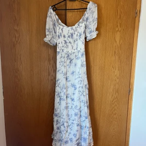 Vit och blå blommig långklänning - Fin långklänning i vitt med blått blommönster från Gina. Klänningen har korta puffärmar och en öppen rygg med knytning. Perfekt för midsommar eller studentmottagningar!