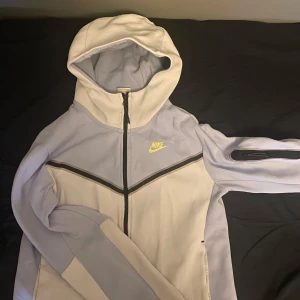 Vit och ljusgrå hoodie från Nike - Säljer en snygg vit och ljusgrå hoodie från Nike med dragkedja och huva. Perfekt för en sportig look. Den har en diskret Nike-logga i gult på bröstet och svarta detaljer vid dragkedjan. Skriv gärna för bilder och frågor den ser mycket bättre ut när den är på. Den är inte ett dugg sliten bara lite på dragkedjan annars i topp skick hör gärna av er :)