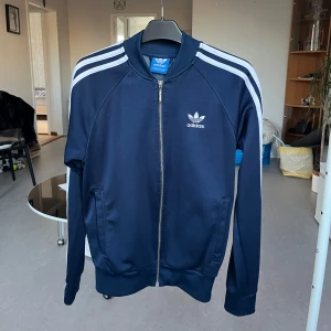 Mörkblå Adidas jacka med dragkedja - Säljer en snygg mörkblå Adidas jacka med dragkedja och de klassiska tre vita ränderna längs ärmarna. Jackan har en broderad Adidas-logga på bröstet och ribbade muddar vid ärmslut och nederkant. Perfekt för en sportig look!