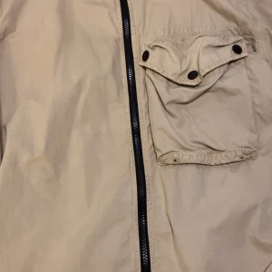 Beige jacka från Stone Island - Säljer en snygg beige jacka från Stone Island med dragkedja och knappdetaljer. Jackan har en praktisk ficka på bröstet och den ikoniska Stone Island-loggan på ärmen. Perfekt för en stilren look.