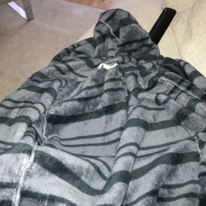 Grå och svart randig morgonrock - Mysig morgonrock i grått och svart med randigt mönster. Perfekt för att mysa i med sin mjuka och bekväma design. Har en praktisk huva och en stor ficka framtill.