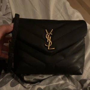 Svart quiltad axelväska från YSL - Snygg svart axelväska från YSL i quiltat skinn med guldiga detaljer. Väskan har en elegant YSL-logga på framsidan och en justerbar axelrem. Perfekt för att ge din outfit en lyxig touch. Kvitto finns ej men har dustbag och verifikationskort samt kod i väskan. Köpt begagnad så äkthet är inte garanterad.